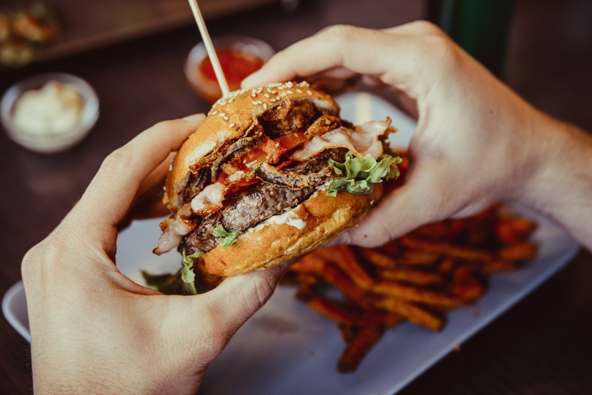 man holds burger with hands and sweet potato fries and dips on background in cafe