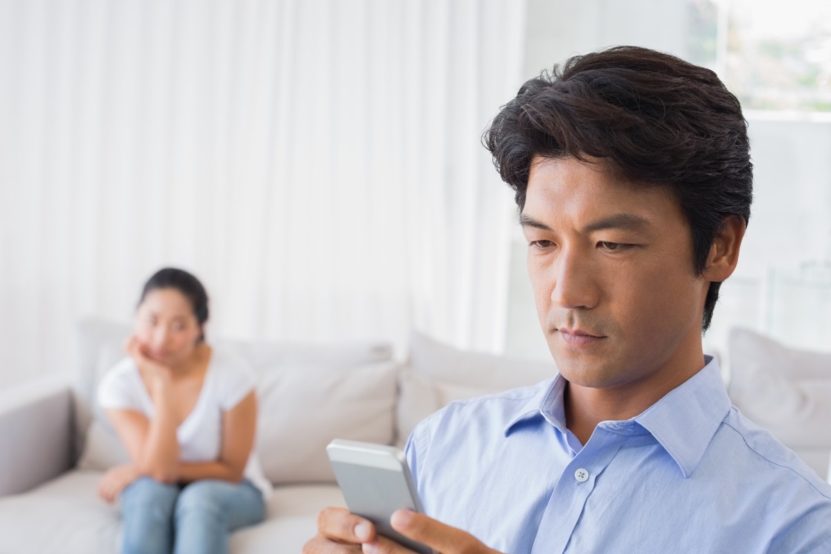 asian man on phone while upset wife looks on from bed