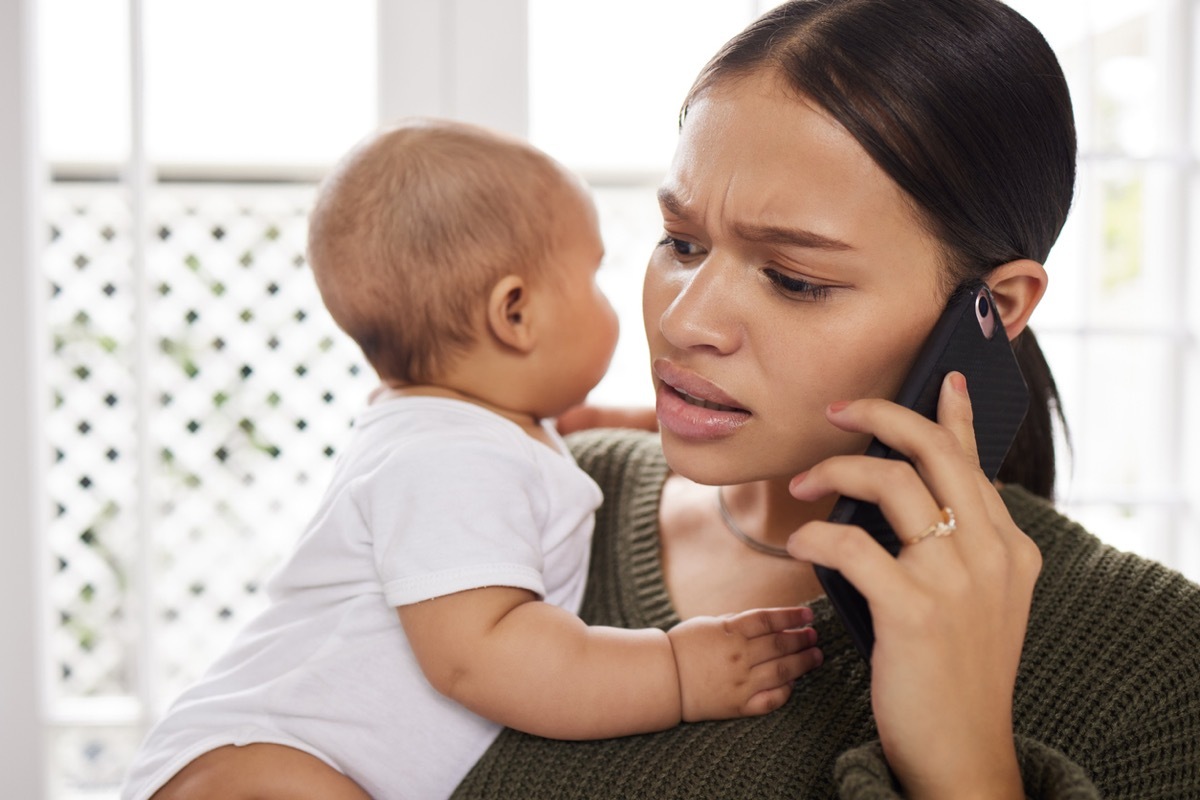 Shot of a young woman using a smartphone while carrying her baby at home