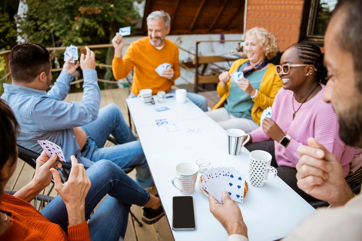 A group of friends of different ages sitting around a patio table playing cards and laughing.