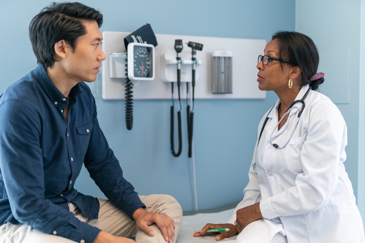 The patient is sitting on an examination table in a clinic. The man's doctor is sitting on the table next to him. The two individuals are facing one another. The patient listens attentively to his doctor's advice.