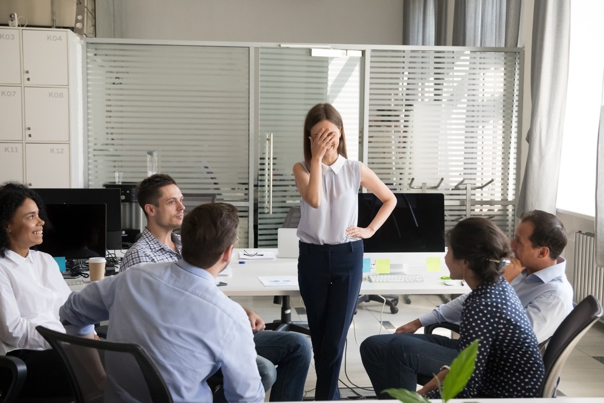 Shy nervous bashful female employee feels embarrassed blushing afraid of public speaking at corporate group team meeting, timid stressed woman hiding face during awkward moment reporting in office (Shy nervous bashful female employee feels embarrassed