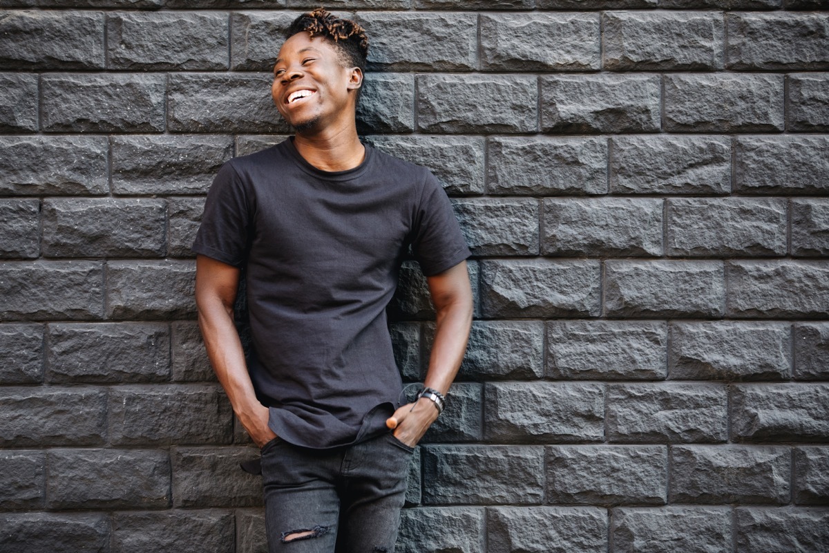 man wearing a black t shirt and black skinny jeans leaning against a gray brick wall, things you shouldn't say about someone's body