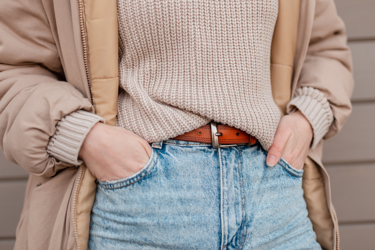 Beautiful stylish woman in fashionable clothes look in a beige Down-padded coat jacket, a knitted vintage Pullover and blue classic jeans stands near a wooden wall.