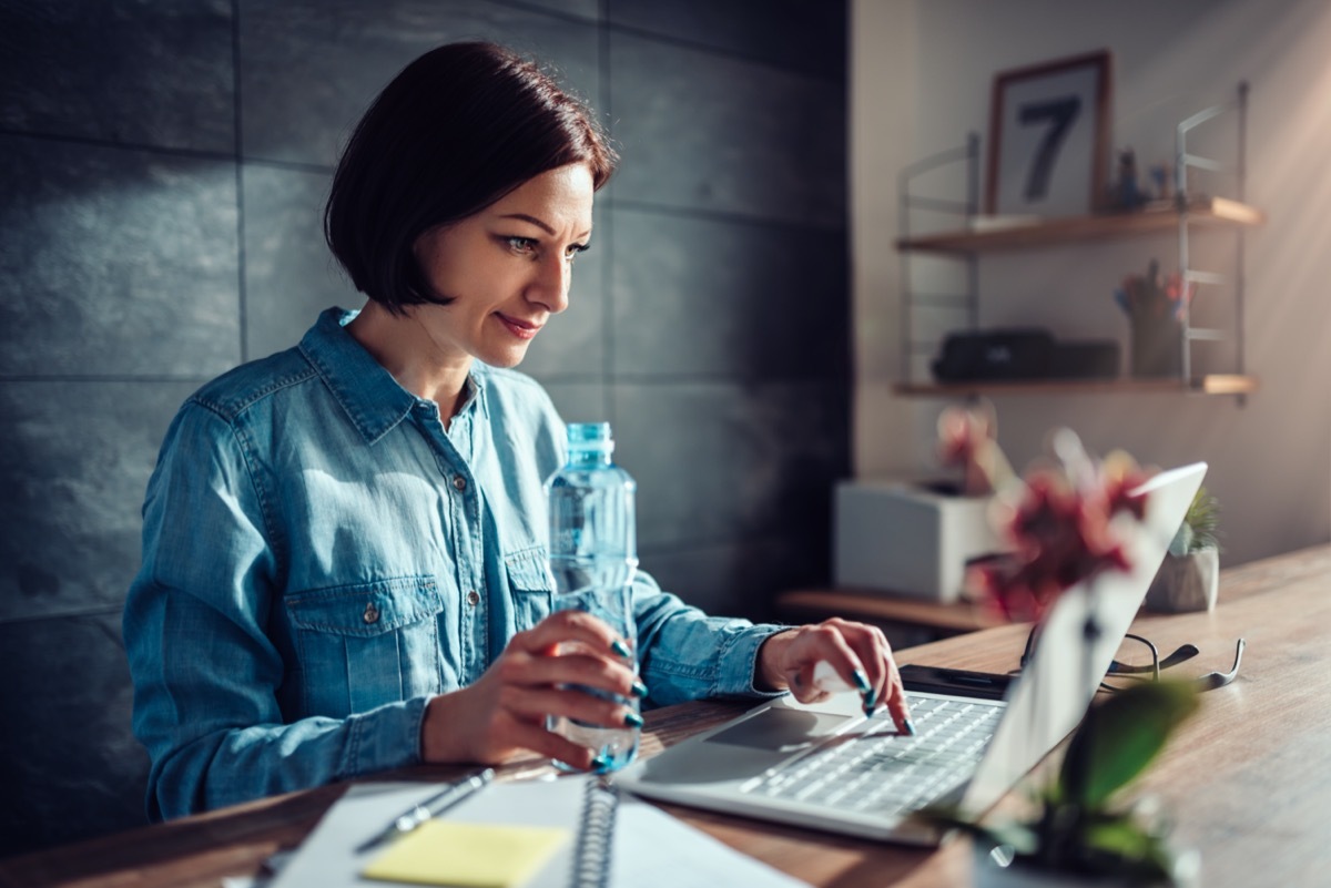 Woman wearing denim shirt using laptop in the office and holding plastic bottle of water