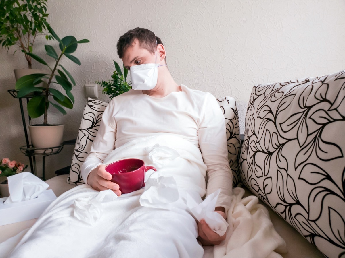 Man in a face mask bowing his head while lying on a sofa at home during quarantine with red cup in his hands