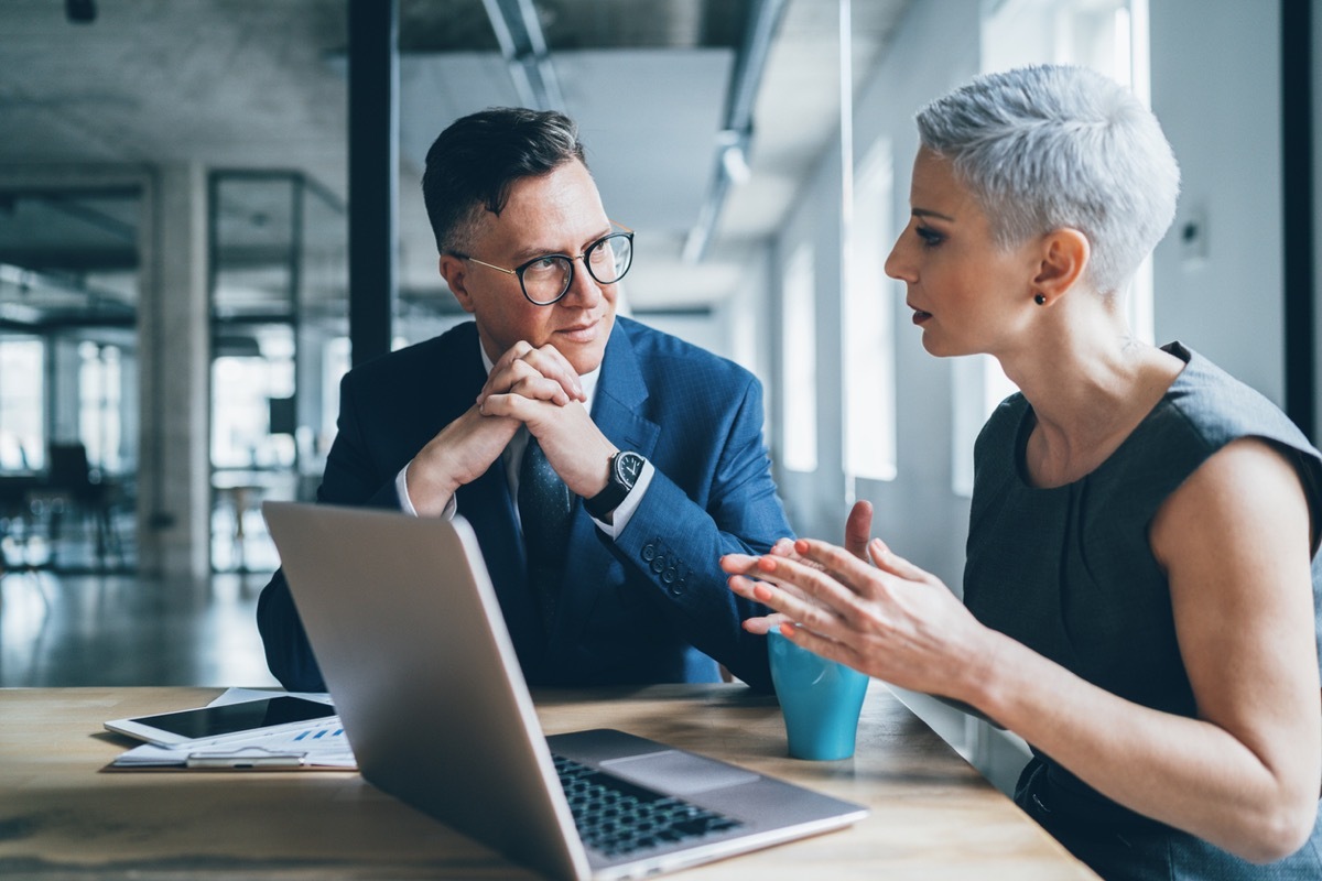 middle aged white man and middle aged white woman talking over a laptop in an office