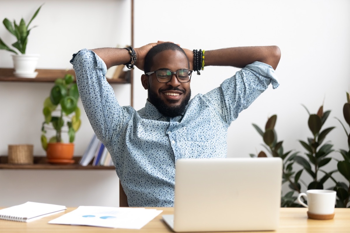 Afro-American businessman holding hands behind head sitting at office desk behind laptop