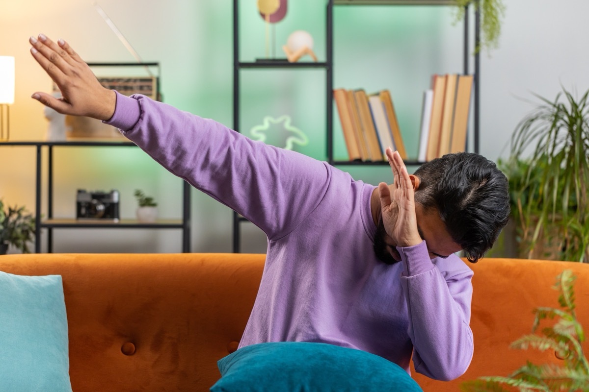 Trendy cheerful positive young indian man having fun dancing and moving to rhythm, dabbing raising hands, making dub dance gesture. Portrait of hindu guy at modern home apartment living room on sofa