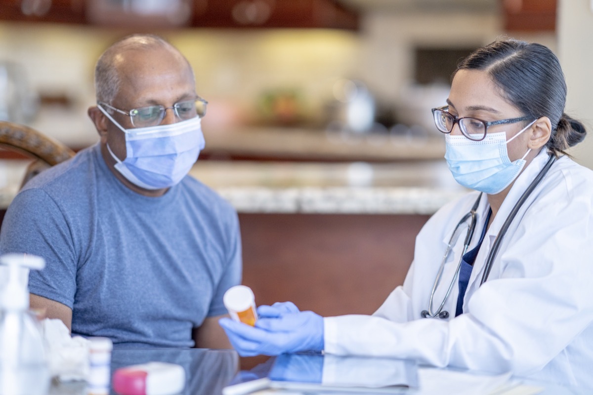 A female doctor is holding a prescription bottle that belongs to her patient. She is reviewing the medicinal ingredients as his patient is seated next to her. Both patient and doctor is using a face mask to help prevent the spread of germs.