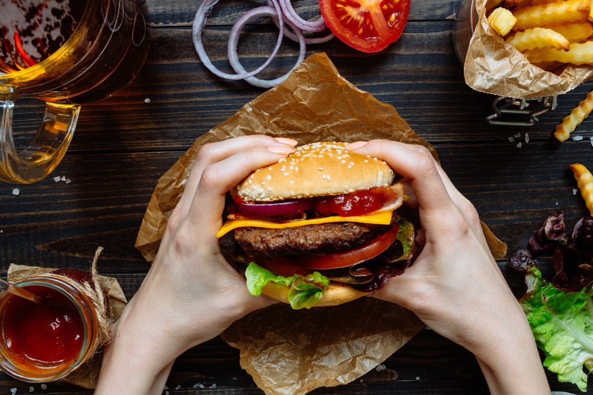 Hands holding fresh delicious burgers with french fries, sauce and beer on the wooden table top view.