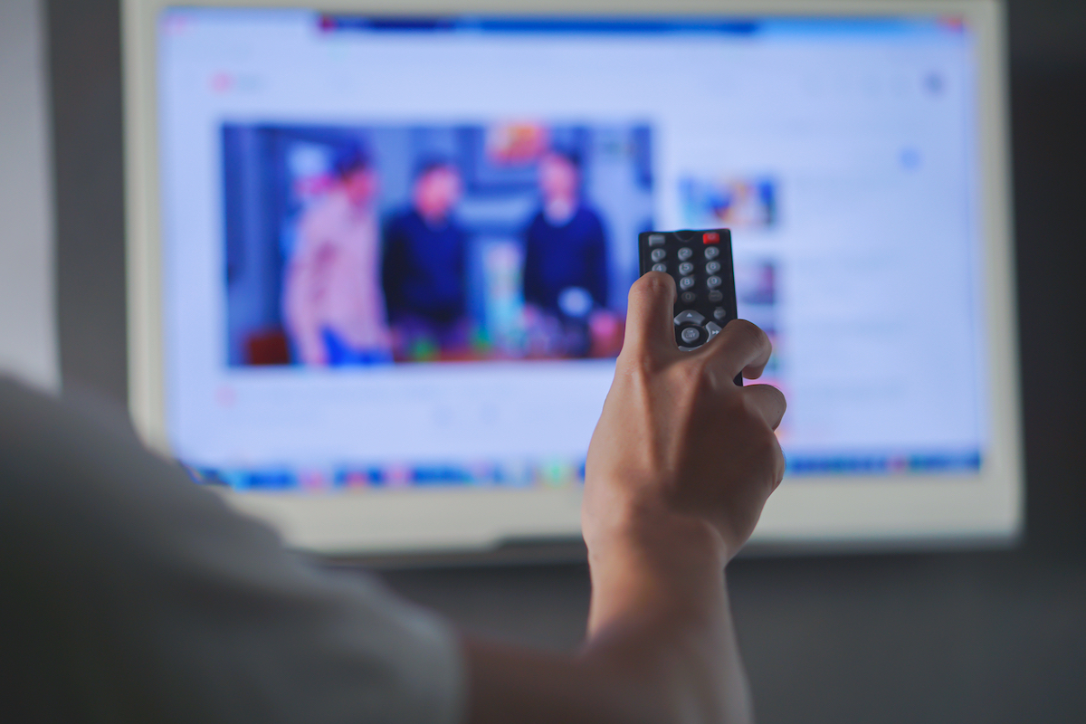 Close up of a remote in a man's hand with a blurry television in the background.