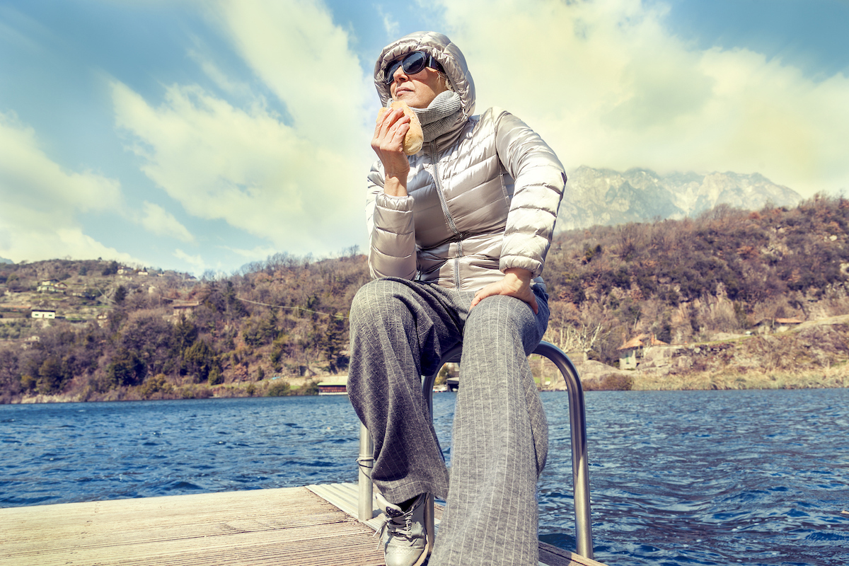 mature woman in puffer coat eating a sandwich on a lake