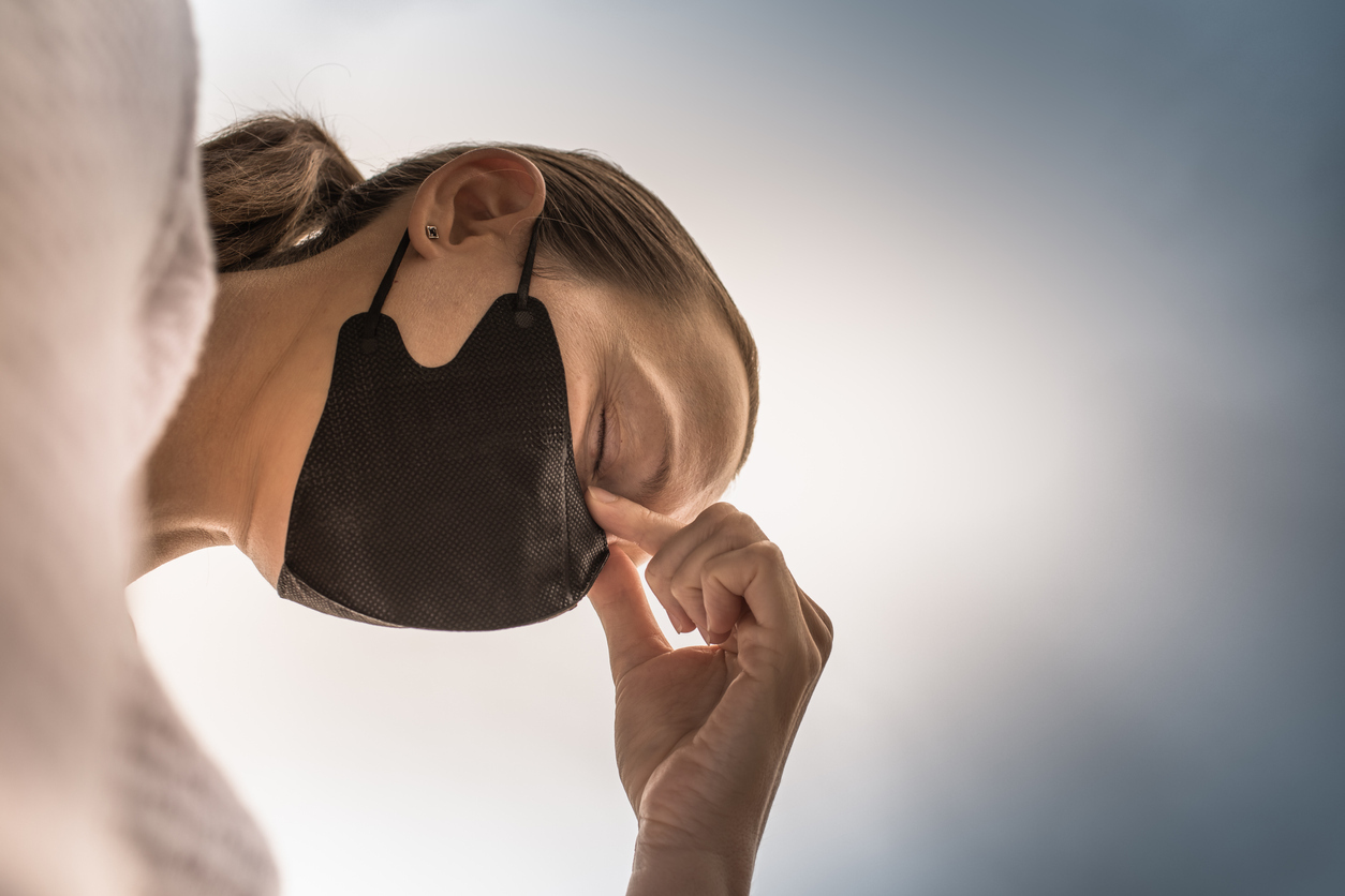 A woman wearing a face mask holds the bridge of her nose with a stressed look on her face