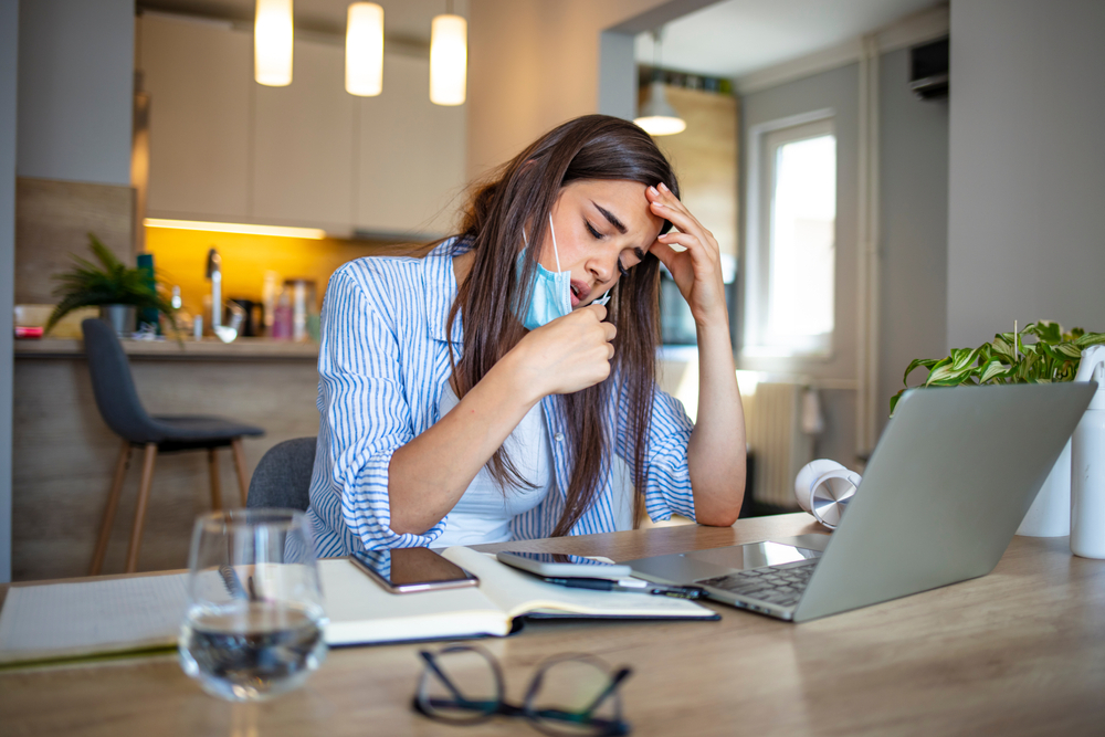 A woman sitting at a laptop holding her head and looking fatigued, perhaps suffering from long COVID