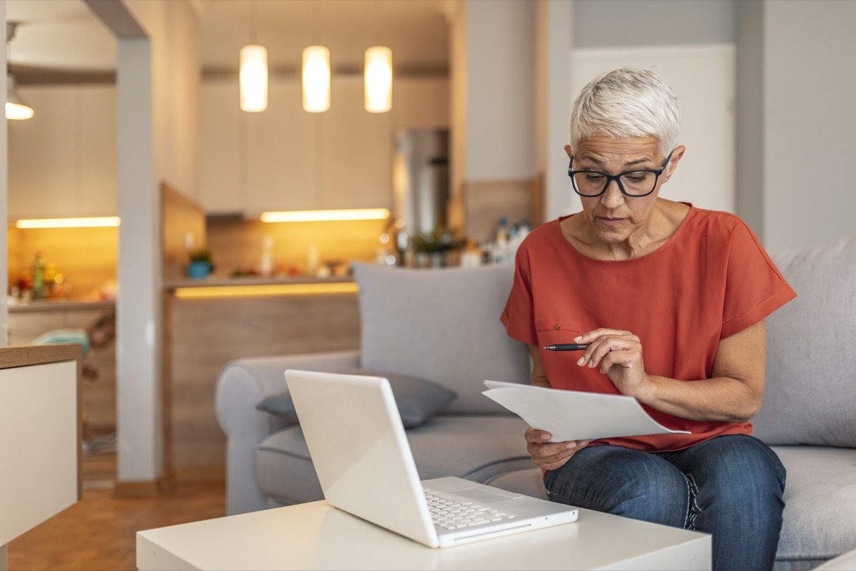 Serious aged woman in eyeglasses checking all bills, calculating expenses. Mature housewife sitting at table with laptop, looking through financial papers with focused expression. Serious woman focused on finding information on paper. Sitting at home in front of a laptop. The concept of business and online student learning.