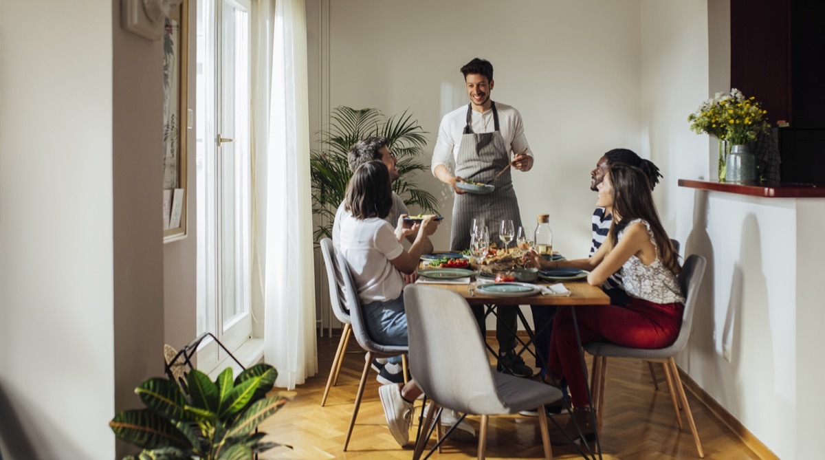 Handsome young man wearing kitchen apron hosting dinner party and serving food to his friends at home.