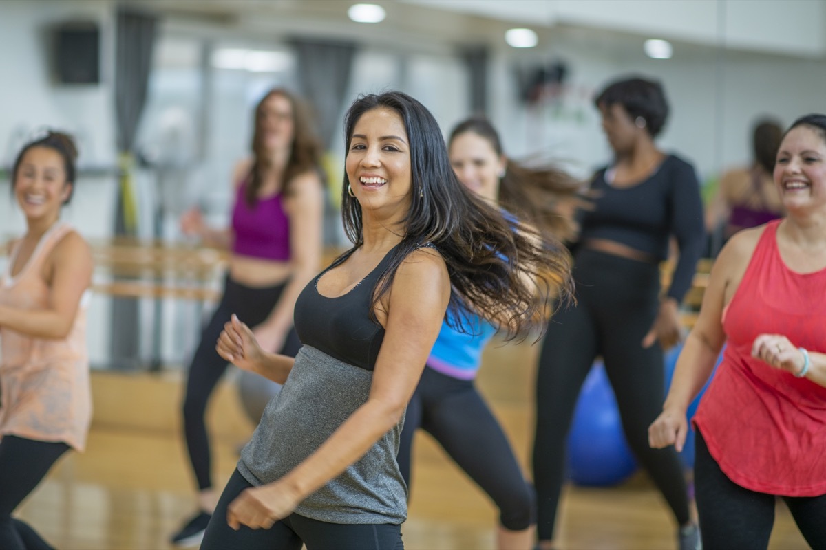 A group of adult women are dancing in a fitness studio. They are wearing athletic clothes. An Ethnic woman is smiling while dancing in the foreground.