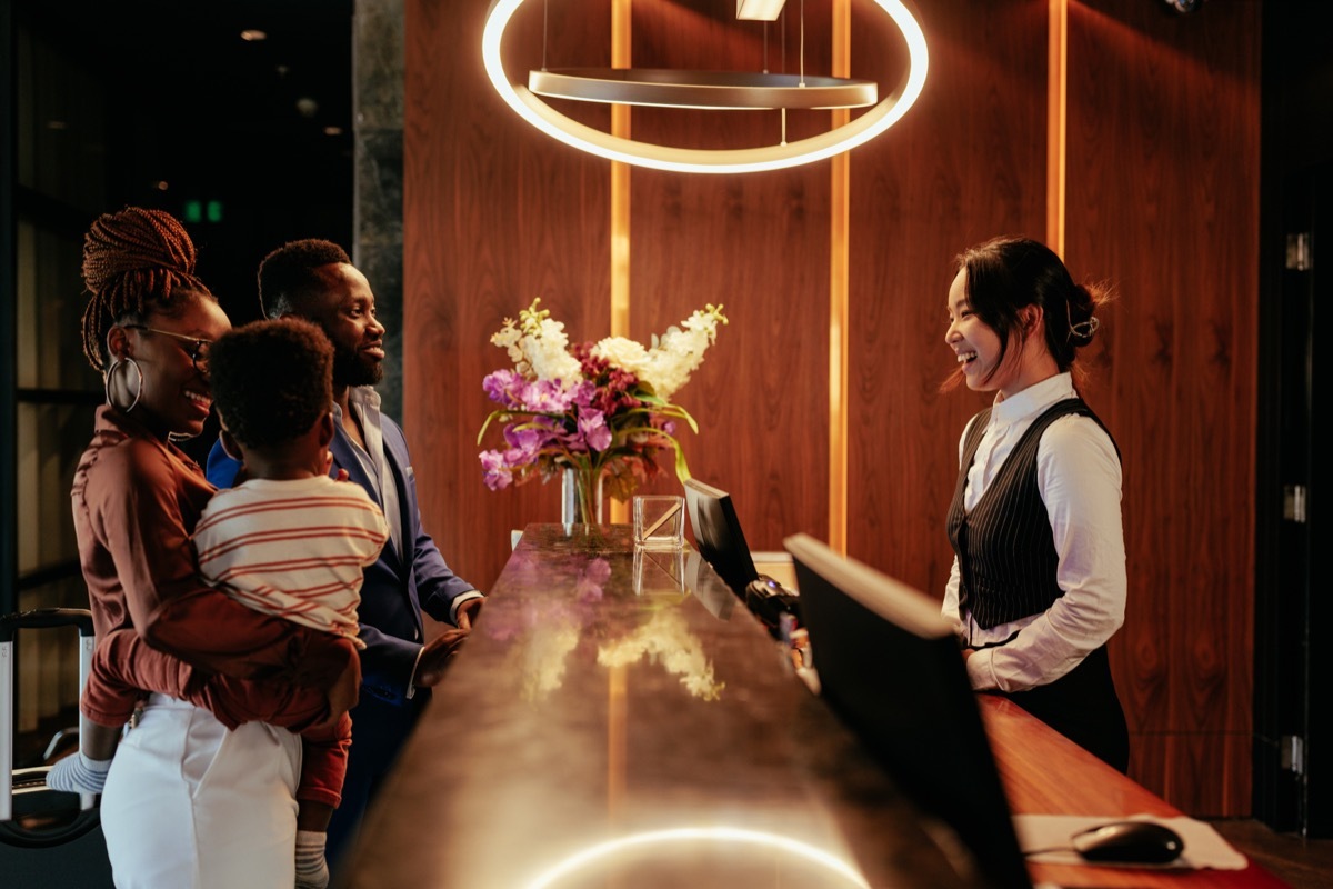 A hotel clerk is welcoming guests that are checking in. It is a wealthy African American couple with their baby boy. The smiling clerk tends to them.