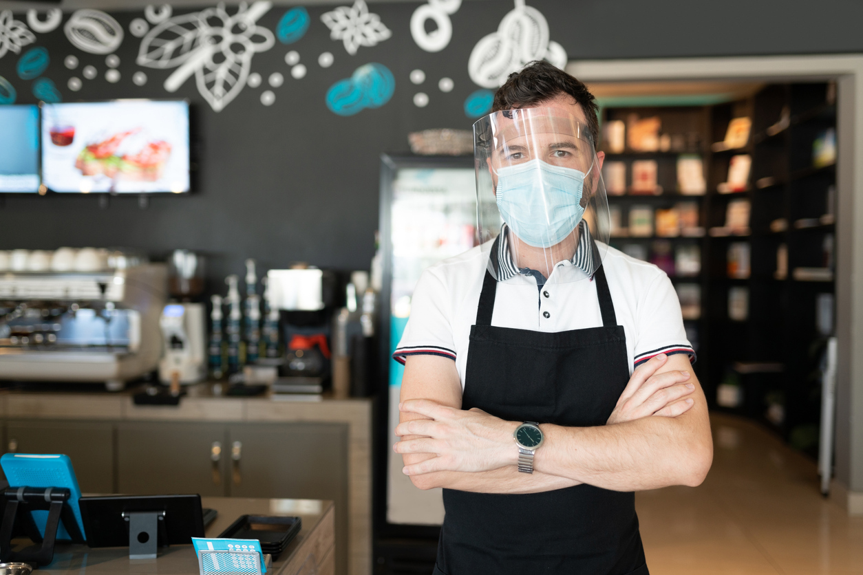 A waiter stands in a cafe with his arms crossed while wearing a face mask and a face shield.