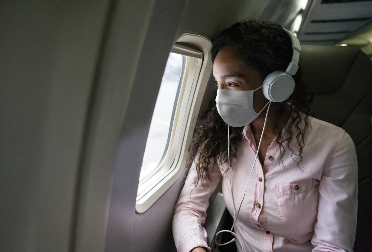 A young woman wearing a mask and headphones looking out the window of an airplane