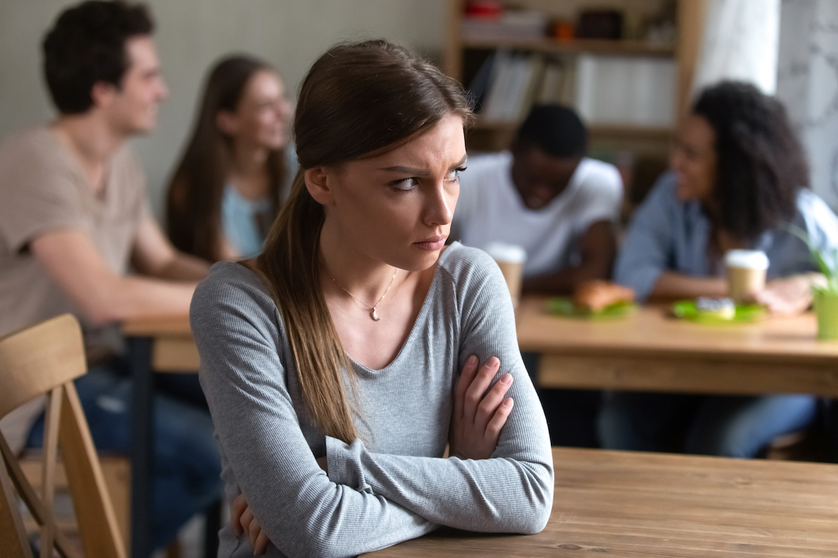 A nervous-looking, antisocial young woman sitting a desk with other people socializing behind her.