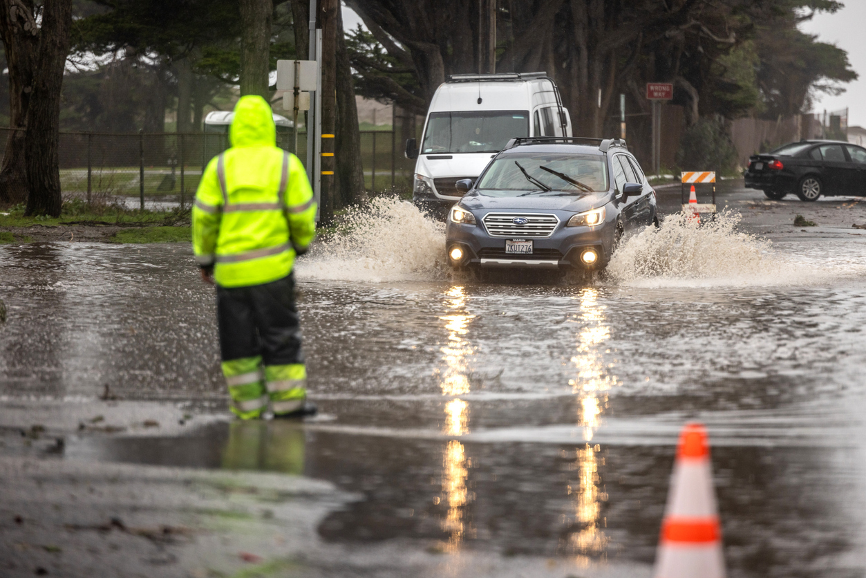 A traffic guard in hi-vis gear guides a car away from a flooded area in a street