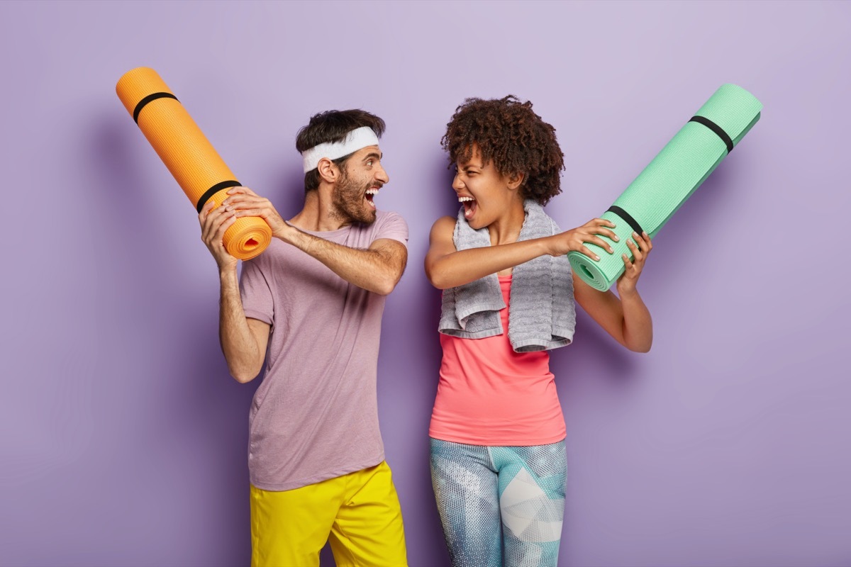 woman and man have fun in gym, fight with rolled up karemats, wear sport clothes, look at each other happily and shout loudly, have regular training together