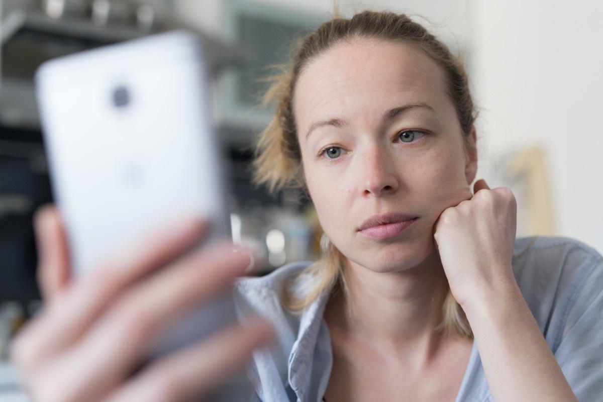 Worried woman indoors at home kitchen using social media apps on phone for video chatting and stying connected with her loved ones