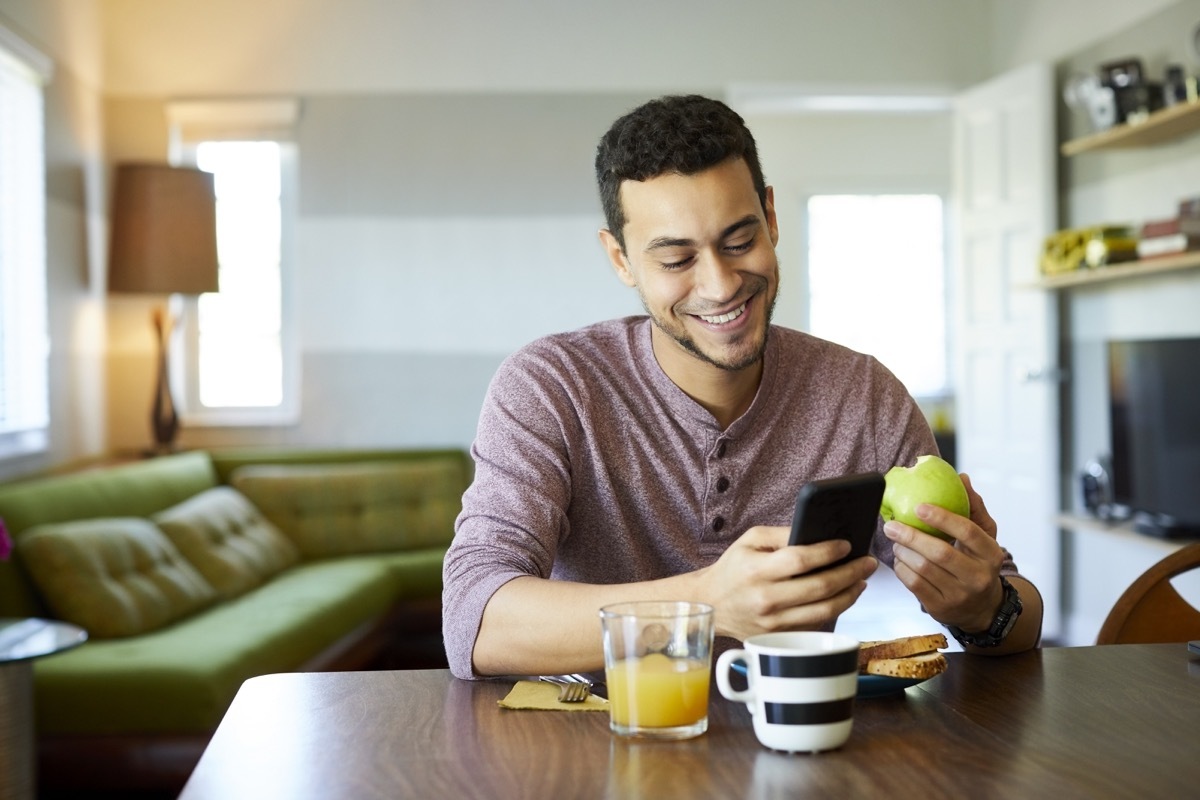 Smiling young man using smart phone at dining table. Male is having granny smith apple at home. He is enjoying social media and breakfast.