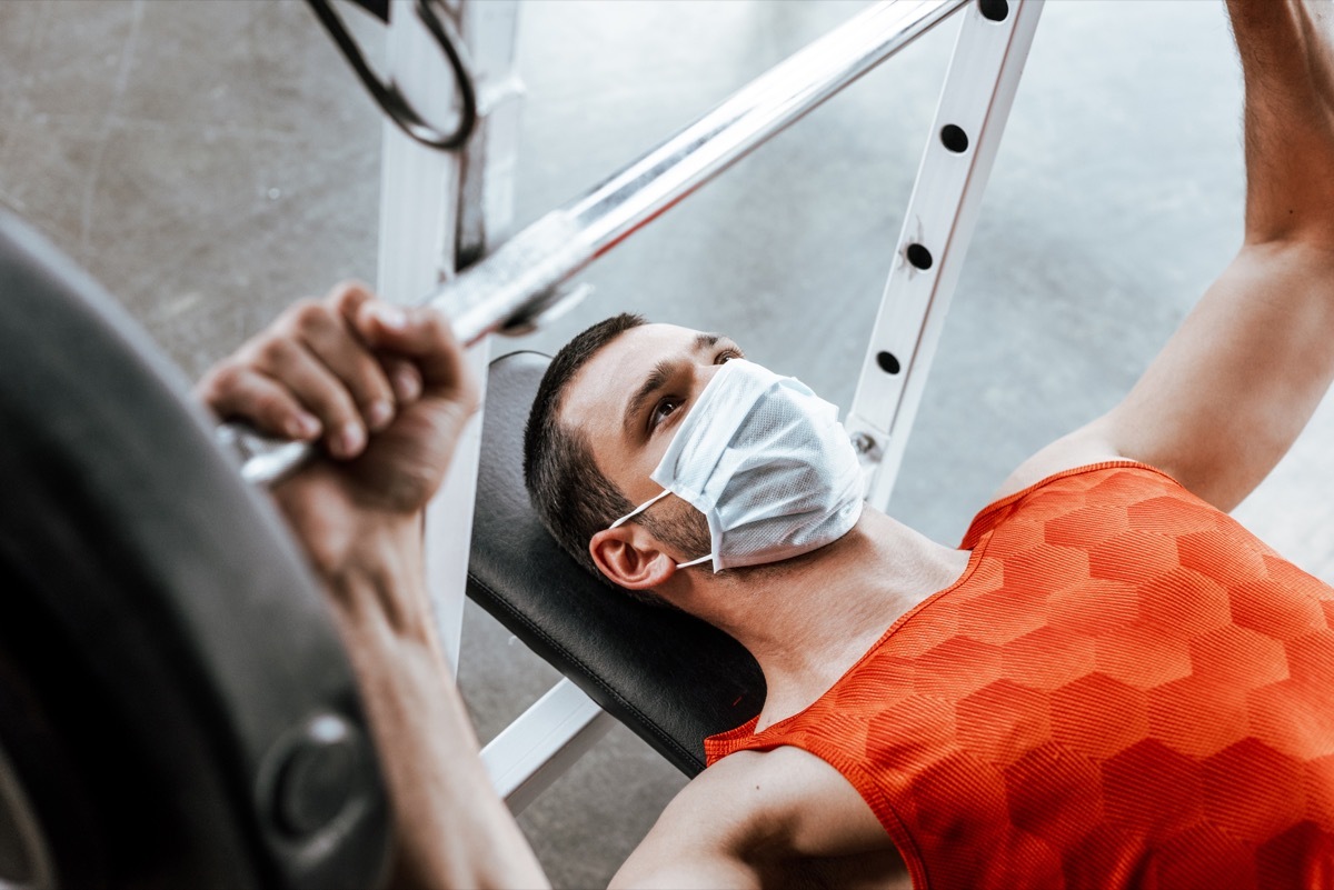 man in medical mask exercising with barbell in gym