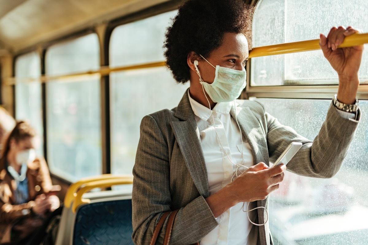 businesswoman with protective face mask using smart phone and looking through the window while commuting by bus.