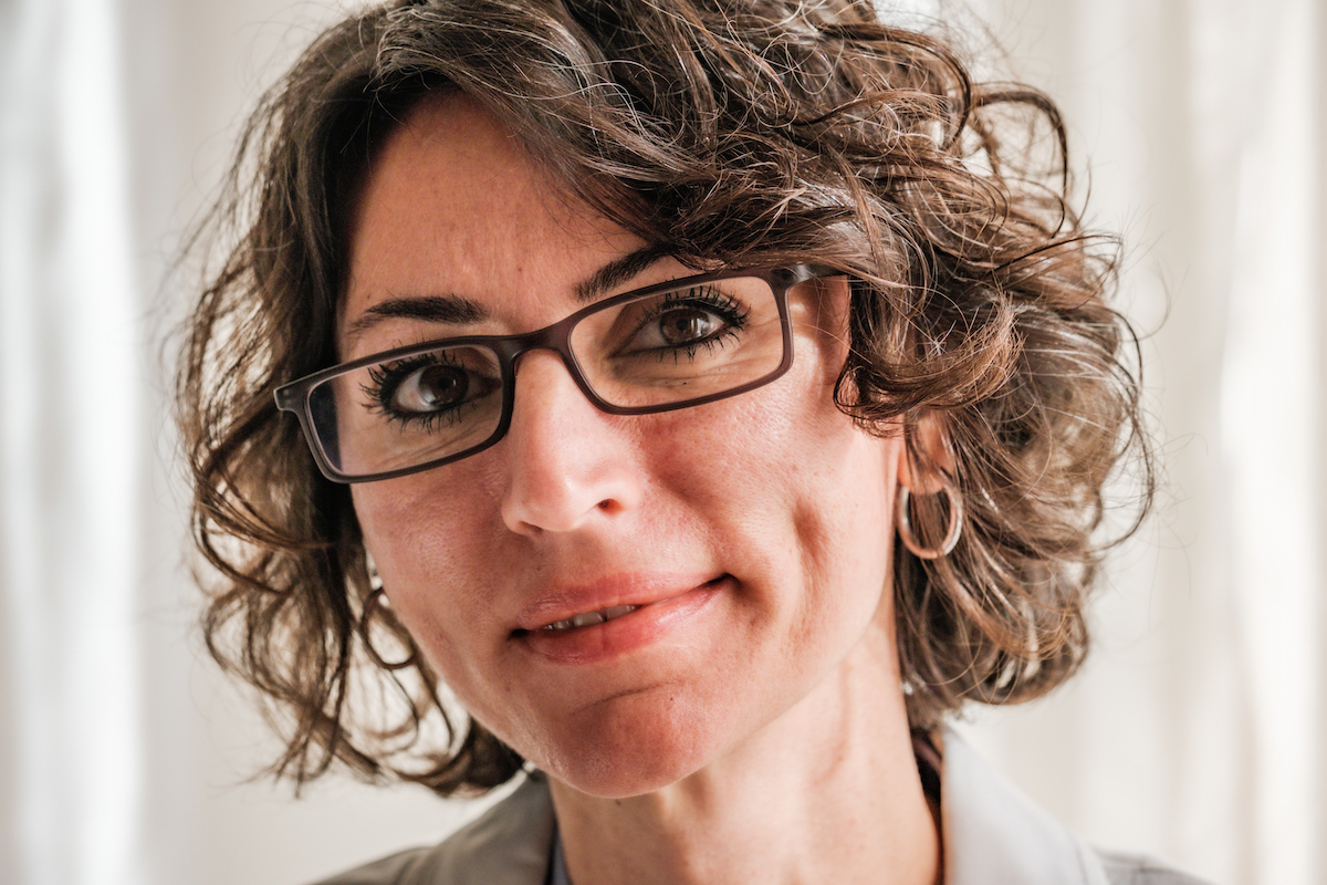 Close up of a middle-aged woman wearing glasses. She has a curly bob haircut, with brown-gray hair