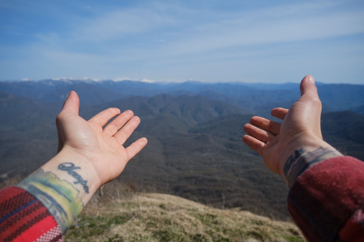 Arms of guy in red shirt and tattoo stretched forward towards mountain