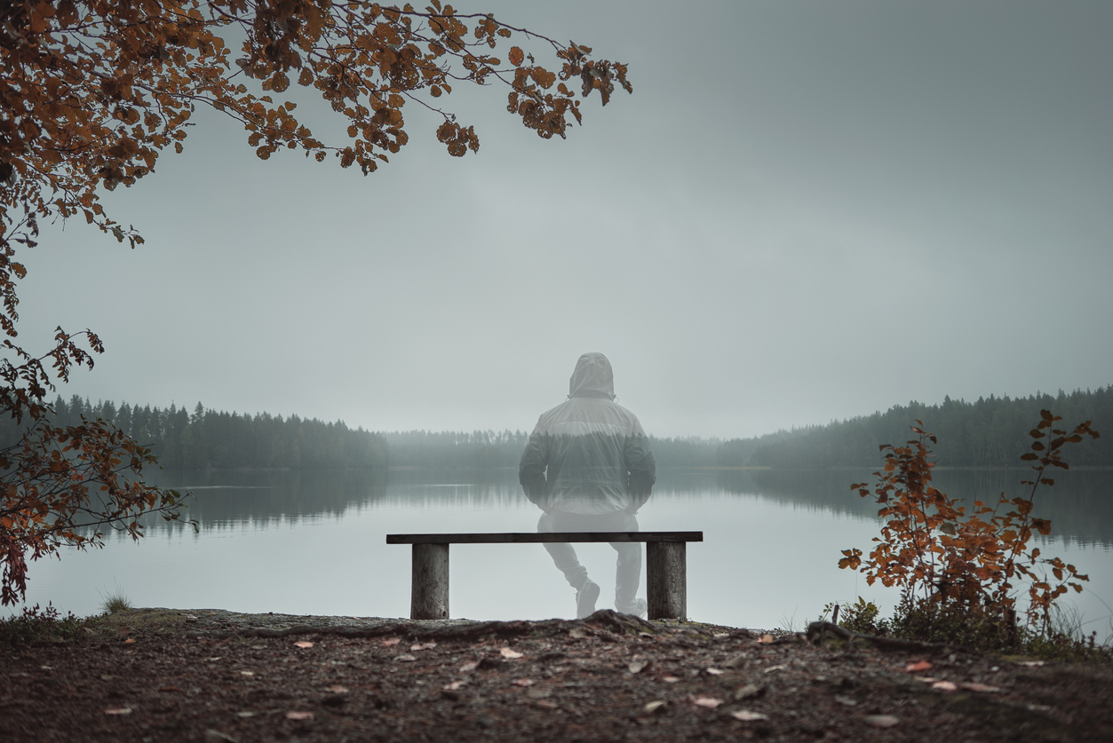 A transparent man is sitting on a bench and looking at the lake