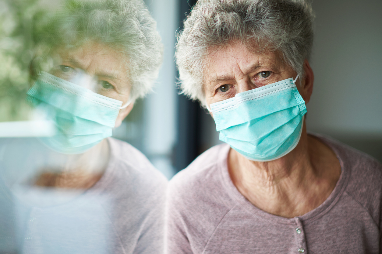 A senior woman wearing a face mask sits at a window with a distressed look on her face.