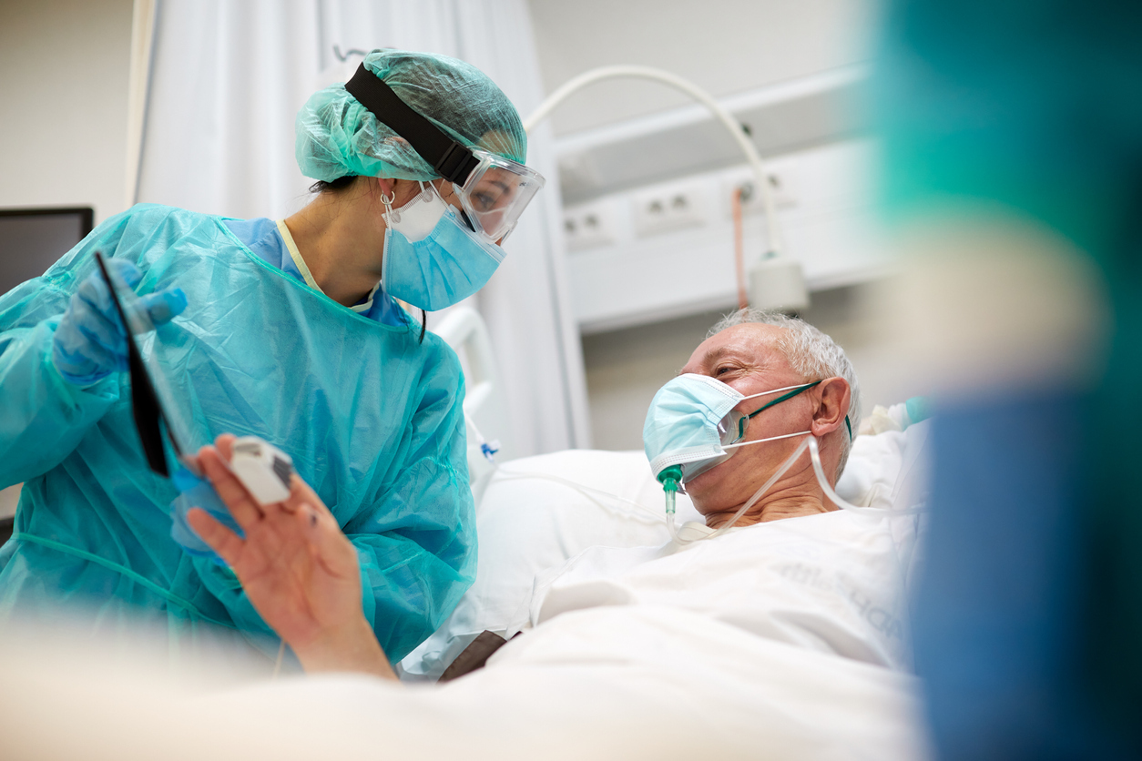 A nurse wearing full protective gear shows a framed photo to an elderly patient in a hospital bed suffering from COVID wearing a face mask and oxygen mask