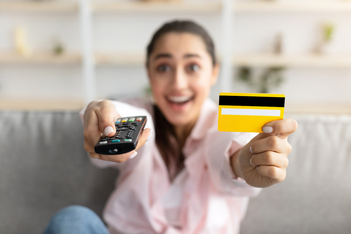 Closeup of excited millennial woman sitting on couch in living room, using tv remote and holding yellow credit card