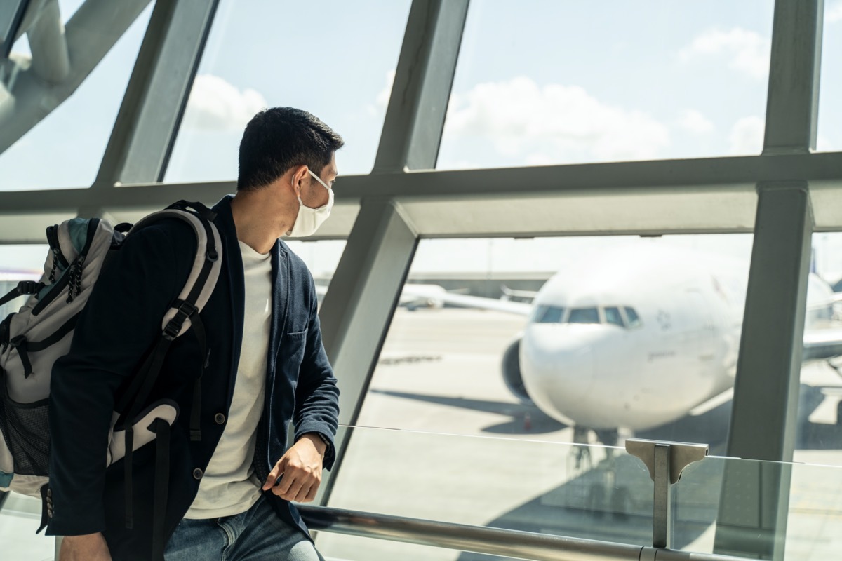 man wears a mask and looks out the airport window at a plane