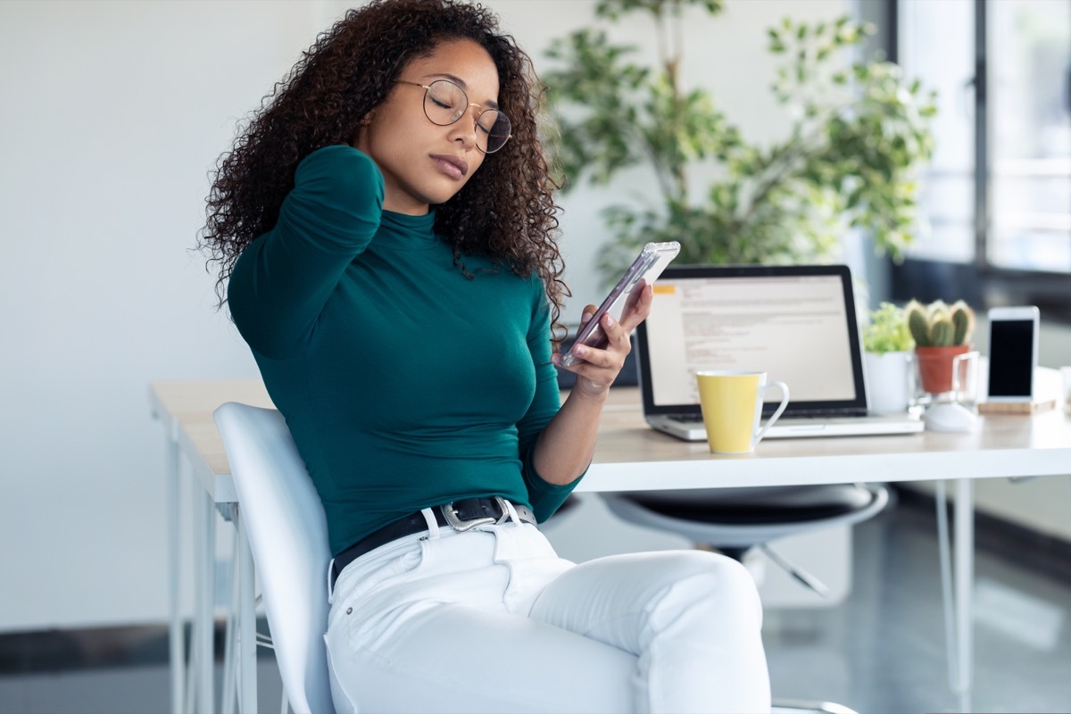 Shot of tired young woman with neck pain holding her mobile phone at the office.