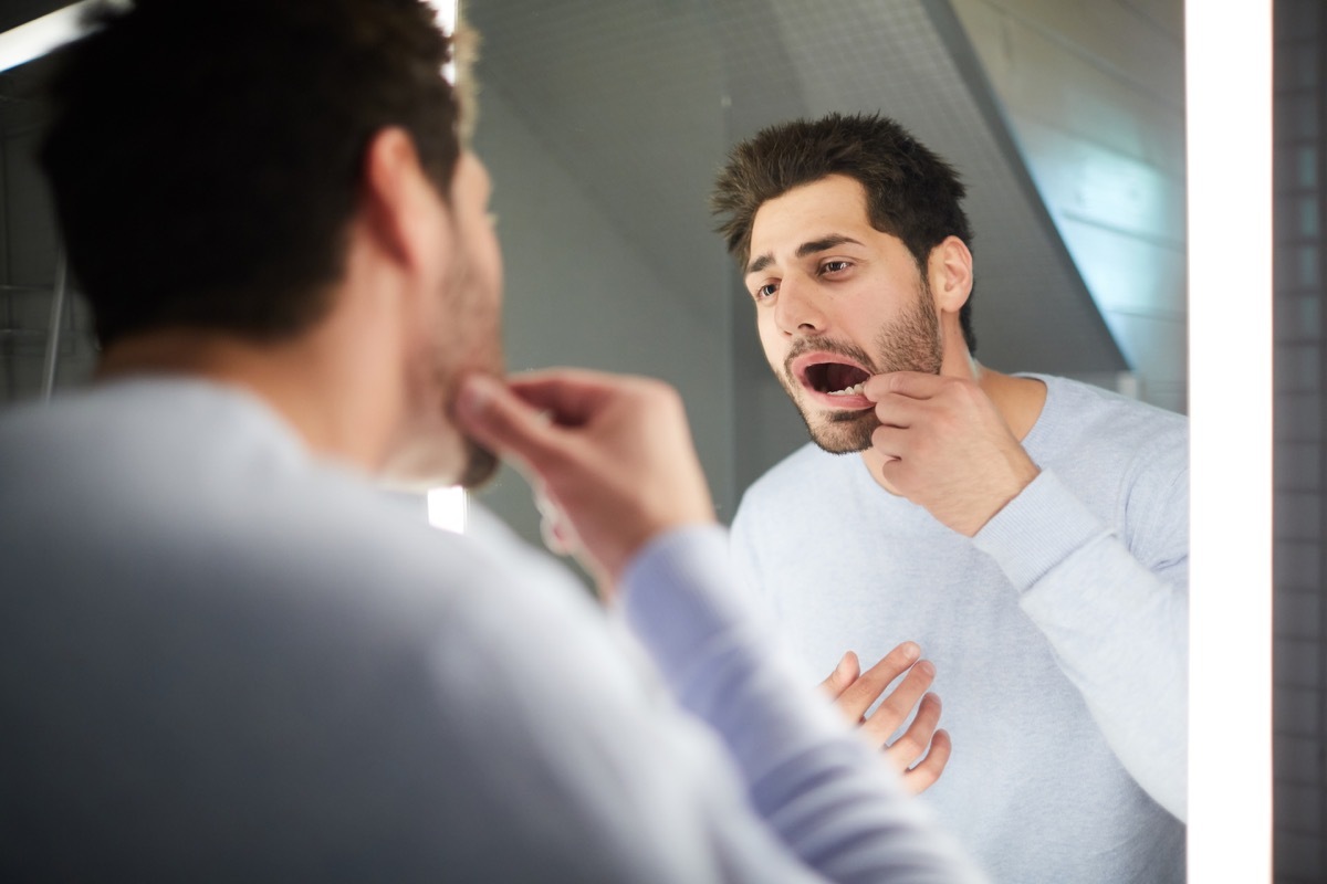 Handsome young man with stubble keeping mouth open while checking tooth and looking into mirror in bathroom