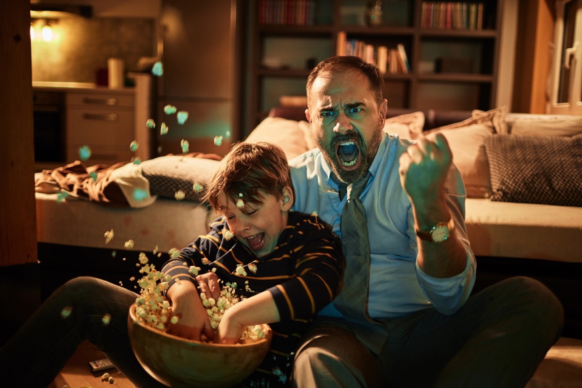 man dressed up excited while watching TV with son throwing popcorn