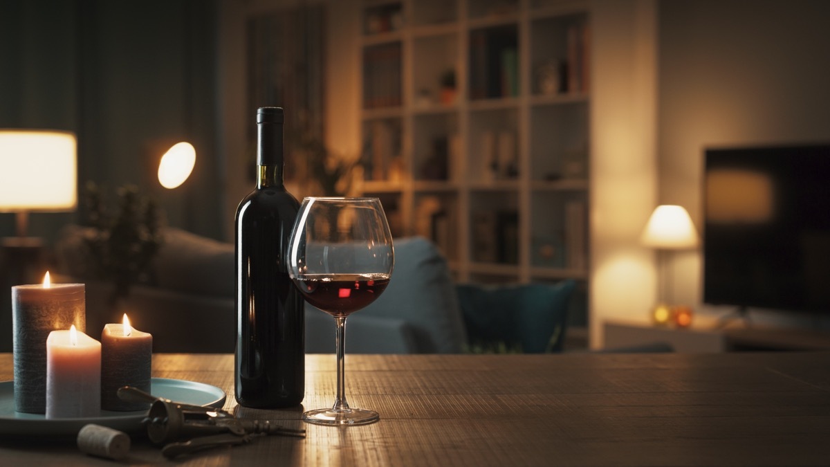 Red wine tasting at home: wine bottle, wineglass, corkscrew and candles on a table in the living room at night