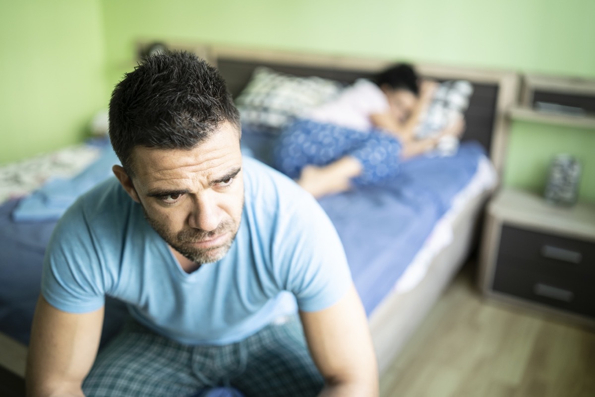 white man looking concerned sitting on the edge of the bed with woman in background