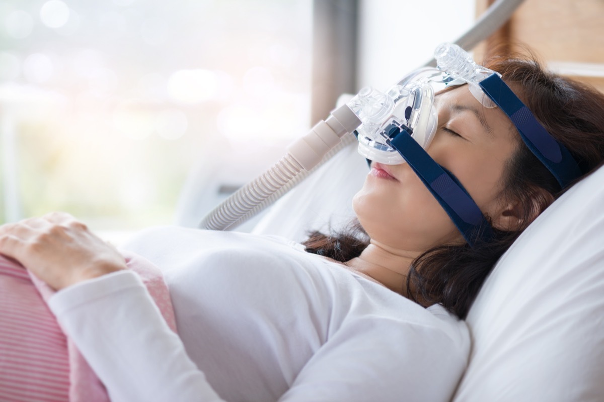 Senior woman using cpap machine to stop choking and snoring from obstructive sleep apnea with bokeh and morning light background. Woman and cpap mask, healthcare concept.