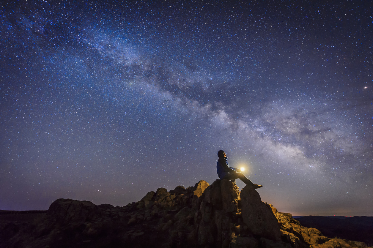 A person sitting on top of a rock outcropping with a light in their hands while looking up at the Milky Way and stars in the night sky