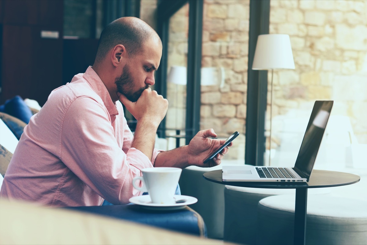 Male student work on his computer while chatting on smart phone, young business man use laptop sitting at sofa of modern coffee shop or hotel hall, freelancer working on notebook at hipster loft space