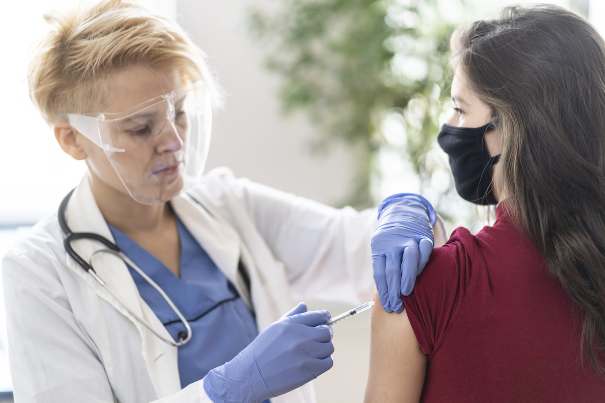 Female doctor vaccinating young woman in her office, wearing a protective face shield