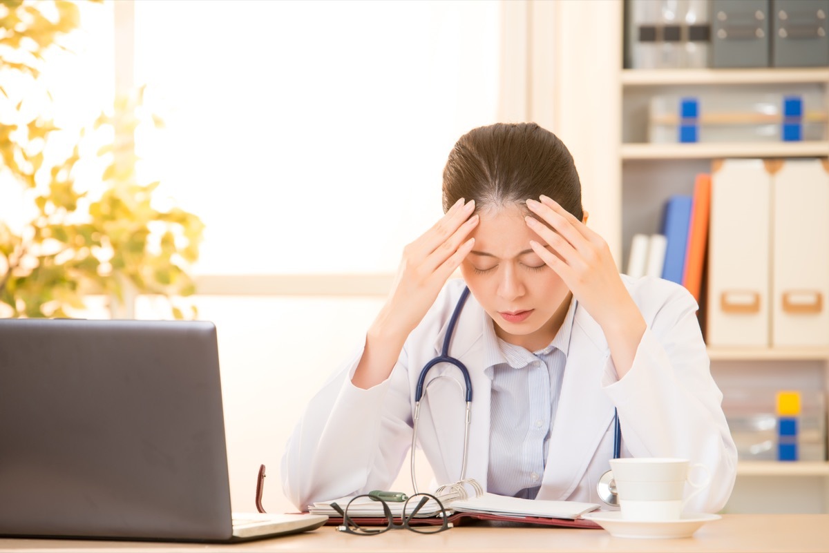 Woman doctor stressed with migraine headache overworked. Health care professional in lab coat wearing stethoscope at hospital