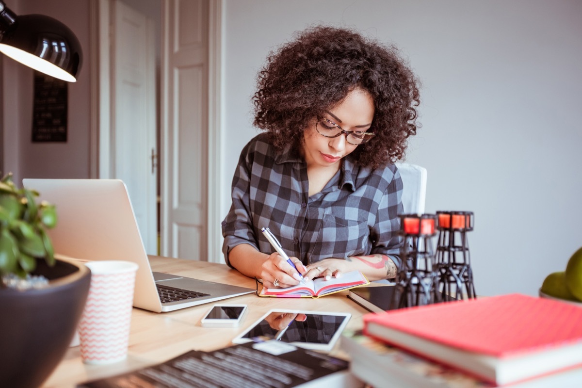 Shot of afro american woman in a home office taking notes in diary. Businesswoman sitting at table and working from home office.